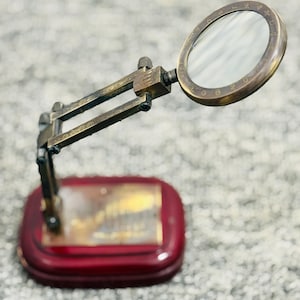 May include: A vintage-style magnifying glass with a brass frame and a round lens. The magnifying glass is mounted on an adjustable arm and a red wooden base. The lens is surrounded by a ring with engraved numbers. The base has a gold-colored plate.