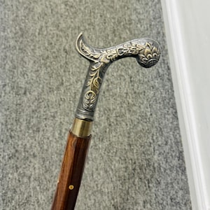 Pode incluir: Bengala vintage com uma pega curva. A pega é prateada com detalhes florais ornamentados. O veio é de madeira castanha escura com uma gola de latão e um pequeno pino dourado.