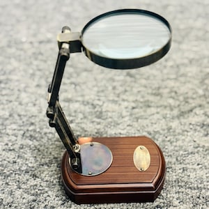 May include: A vintage-style magnifying glass with a dark wood base and adjustable metal arm. The round magnifying lens is set in a brass-colored frame. The base has a brass-colored circular accent and an oval plaque.