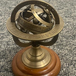 Peut inclure: Une sphère armillaire en laiton avec des symboles du zodiaque et des graduations, montée sur une base en bois. La sphère présente des anneaux entrecroisés et un globe central. La base est une plateforme en bois polie et ronde. C'est un objet décoratif.