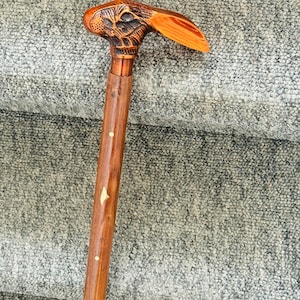 Puede incluir: Bastón de madera con un mango tallado en forma de cabeza de conejo. El mango es marrón y naranja, con detalles intrincados. El eje es de color marrón pulido con incrustaciones blancas. Un accesorio elegante y funcional.