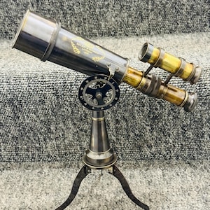 Puede incluir: Un telescopio de latón antiguo con un trípode. El telescopio tiene un acabado oscuro con detalles dorados y el texto "KELVIN & HUGHES LONDON". El soporte tiene tres patas y un dial de ajuste circular.