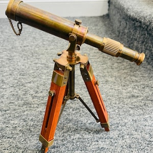 Könnte beinhalten: Ein antikes Messing-Teleskop mit einem Holzstativ. Das Teleskop hat einen langen Lauf mit Kette und geriffeltem Okular. Die Stativbeine bestehen aus rotbraunem Holz und Messingbeschlägen. Ein klassisches nautisches oder wissenschaftliches Instrument.