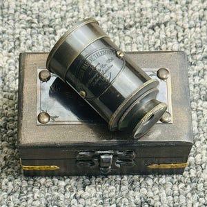 Puede incluir: Un telescopio de latón antiguo y una caja de madera. El telescopio es negro con detalles y texto. La caja es de color marrón oscuro con detalles en latón y un cierre.