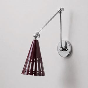 Può includere: Una lampada da parete con finitura in cromo lucido e braccio articolato regolabile. Il paralume conico è di un colore bordeaux intenso con una serie di intagli verticali e perforazioni circolari lungo il bordo inferiore della superficie.