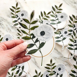 May include: A set of white floral stickers with green leaves and gold accents. The stickers feature a single white flower with a black center, surrounded by leaves and a gold circle.