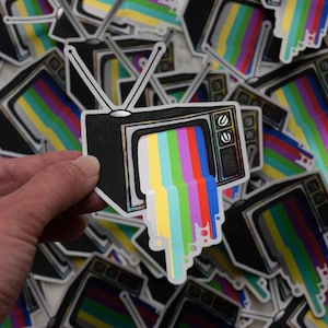 May include: A black and white sticker of a vintage television with rainbow coloured static coming out of the screen. The sticker is being held by a hand.