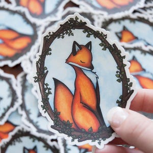 May include: A sticker featuring a fox with a white chest and a bushy tail, sitting in a wreath of leaves. The fox is looking to the side with its eyes closed. The background is a light blue watercolor wash.