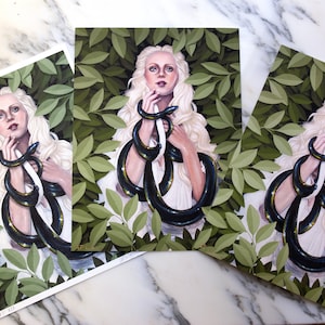 Op de afbeelding: Een vrouw met lang blond haar is omringd door weelderig groen. Ze houdt twee zwarte slangen in haar handen. De slangen zijn om haar armen en nek gewikkeld. De vrouw draagt een witte jurk.