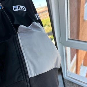 May include: Black and white FILA track jacket with a blue accent on the collar. The jacket features a full zipper and the FILA logo on the chest. The jacket has a white panel on the sleeve and a black ribbed hem.