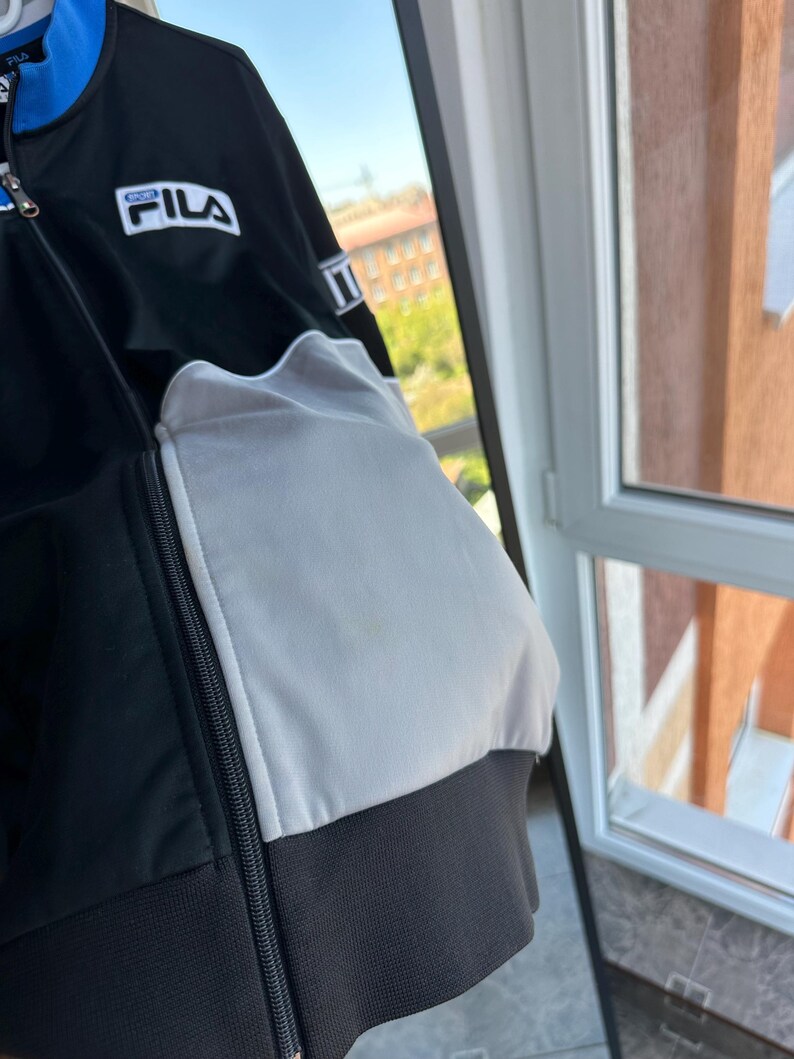 May include: Black and white FILA track jacket with a blue accent on the collar. The jacket features a full zipper and the FILA logo on the chest. The jacket has a white panel on the sleeve and a black ribbed hem.