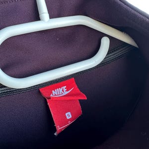 May include: A dark purple Nike track jacket with a black zip and a red Nike label. The label displays the Nike logo and the size "S". The jacket is suspended on a white clothes hanger.