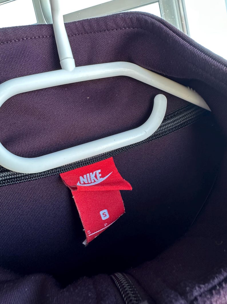 May include: A dark purple Nike track jacket with a black zip and a red Nike label. The label displays the Nike logo and the size "S". The jacket is suspended on a white clothes hanger.