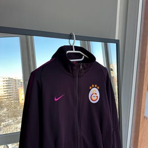 May include: A dark purple track jacket with a high collar and a zip closure. The jacket features a pink Nike swoosh logo and a circular emblem with a star and crescent design. The jacket is hanging on a white hanger.