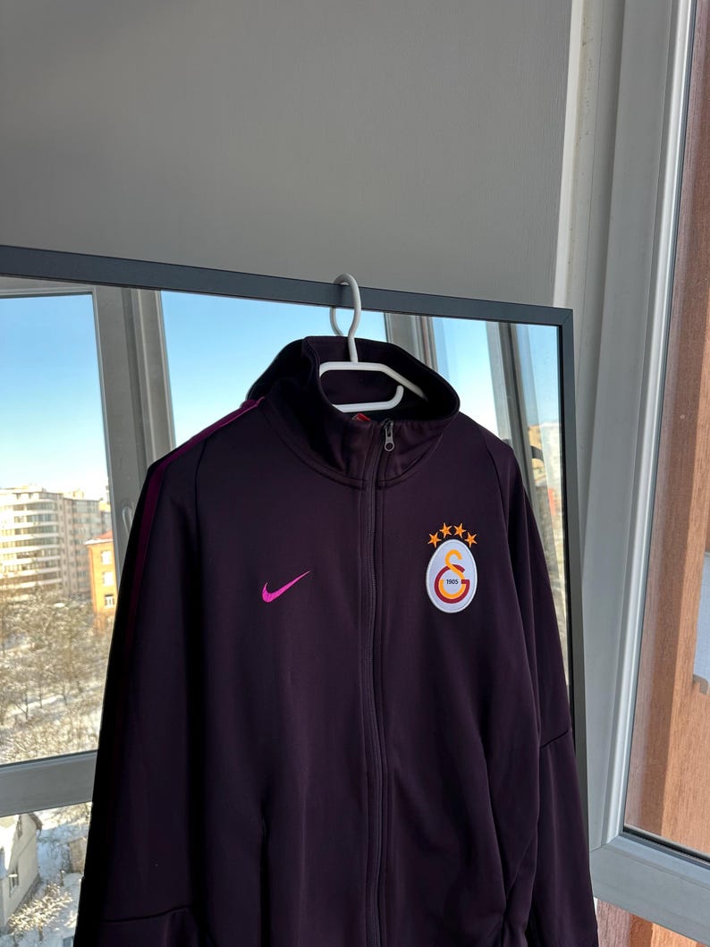 May include: A dark purple track jacket with a high collar and a zip closure. The jacket features a pink Nike swoosh logo and a circular emblem with a star and crescent design. The jacket is hanging on a white hanger.