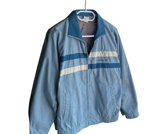 May include: Light blue Adidas track jacket with a full zipper closure. The jacket features a light blue collar and two horizontal stripes across the chest: a light blue stripe and a white stripe. The Adidas logo is visible on the chest.