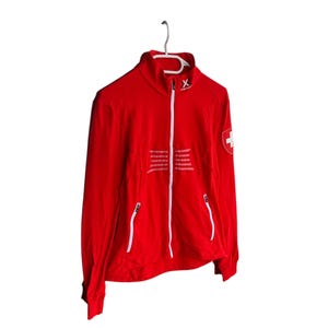 May include: A vibrant red zip-up jacket with a high collar, white zips, and a Swiss flag patch on the upper left sleeve. The jacket features white embroidered stripes on the front and is suspended from a white hanger.