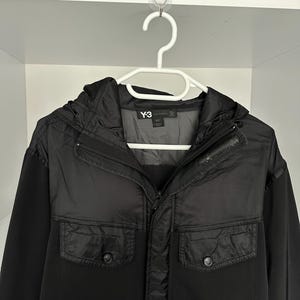 K&ouml;nnte beinhalten: Schwarze Jacke mit Kapuze und zwei Vordertaschen, h&auml;ngt an einem wei&szlig;en Kleiderb&uuml;gel. Das Markenlogo "Y-3" ist auf der Jacke sichtbar.