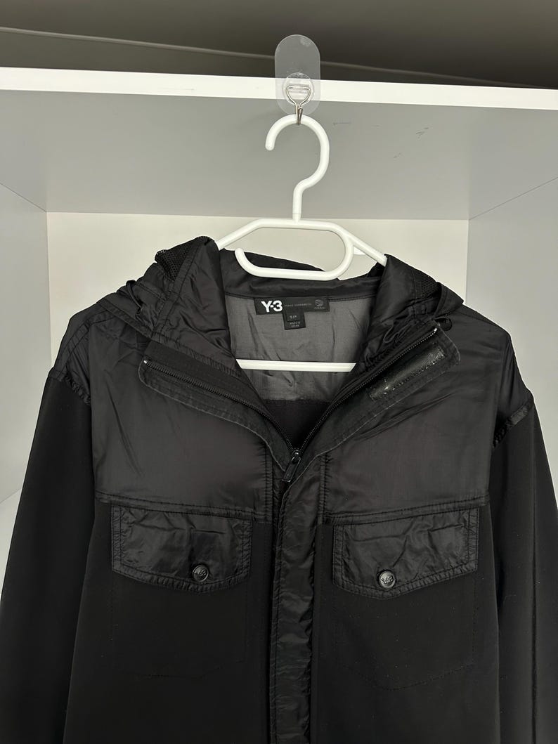 K&ouml;nnte beinhalten: Schwarze Jacke mit Kapuze und zwei Vordertaschen, h&auml;ngt an einem wei&szlig;en Kleiderb&uuml;gel. Das Markenlogo "Y-3" ist auf der Jacke sichtbar.
