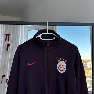 May include: A dark purple zip-up jacket with a high collar, featuring a pink Nike swoosh and a team logo patch. The patch includes a circular emblem with the text "1905" and three gold stars.