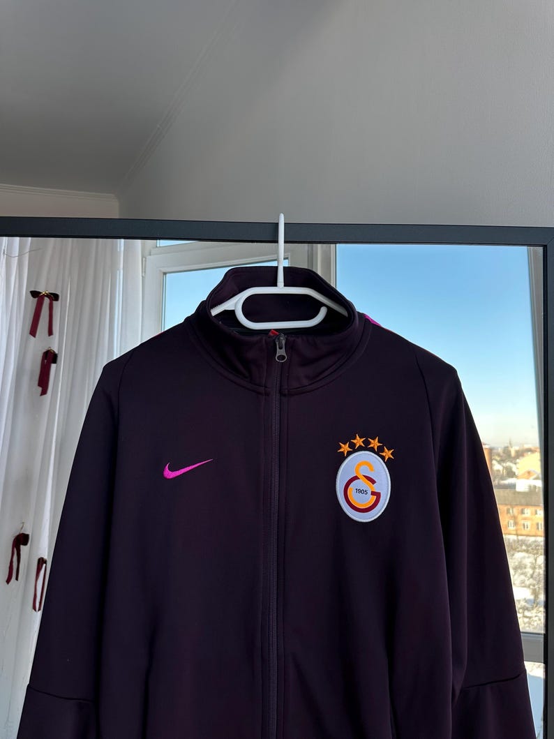 May include: A dark purple zip-up jacket with a high collar, featuring a pink Nike swoosh and a team logo patch. The patch includes a circular emblem with the text "1905" and three gold stars.