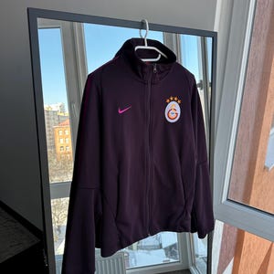 May include: A dark purple zip-up athletic jacket with a high collar, featuring a small pink Nike logo and a team emblem on the chest. The jacket has a burgundy stripe down the sleeves and is hanging on a white hanger.