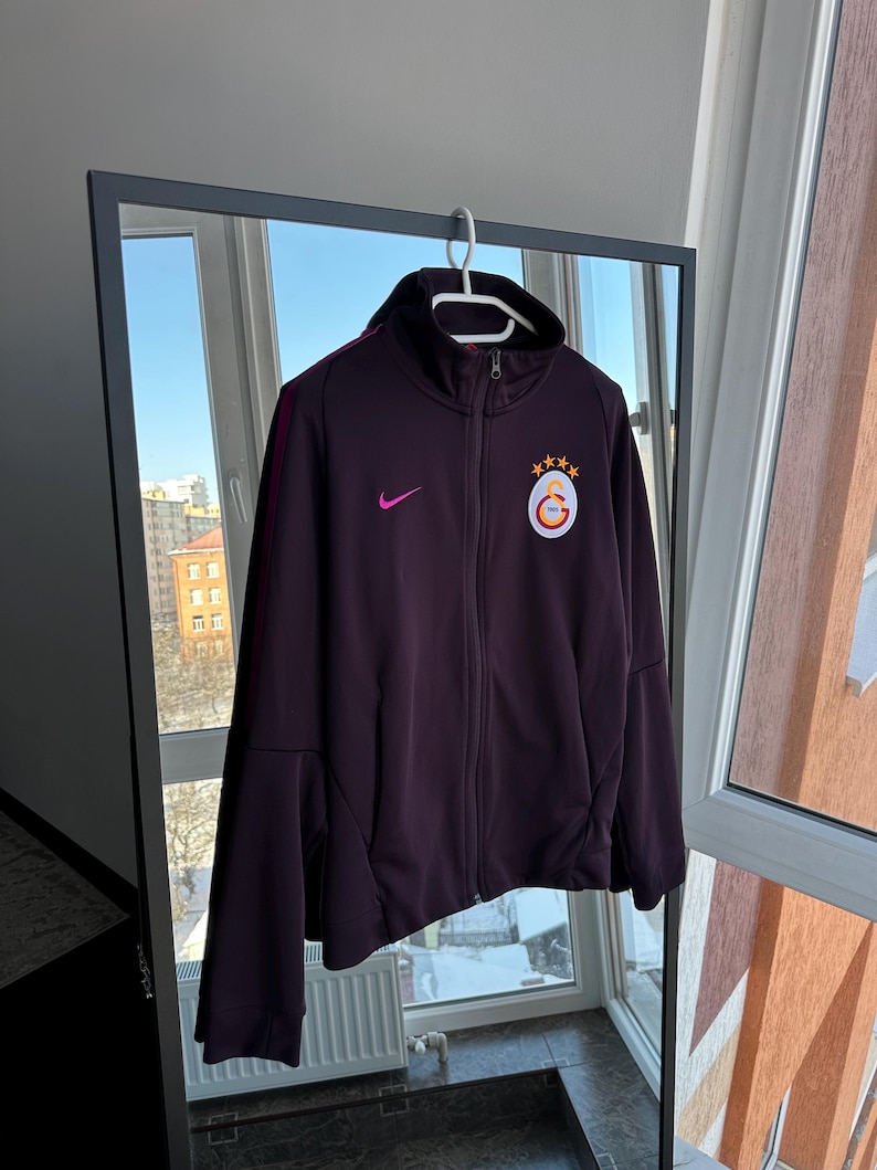 May include: A dark purple zip-up athletic jacket with a high collar, featuring a small pink Nike logo and a team emblem on the chest. The jacket has a burgundy stripe down the sleeves and is hanging on a white hanger.