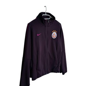May include: A dark purple zip-up jacket with a black collar and a white hanger. The jacket features a pink Nike logo and a team emblem with stars. The jacket has a full-length zipper and side pockets.