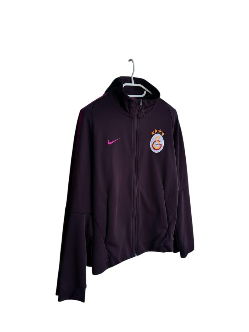 May include: A dark purple zip-up jacket with a black collar and a white hanger. The jacket features a pink Nike logo and a team emblem with stars. The jacket has a full-length zipper and side pockets.