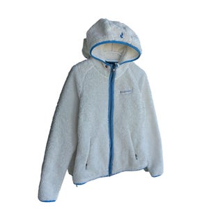 Puede incluir: Chaqueta con capucha de forro polar blanco con cremallera y ribetes azules. La chaqueta tiene una capucha con ribetes azules y un pequeño logotipo en el pecho. Tiene dos bolsillos laterales con cremallera. La chaqueta es de material suave y esponjoso.