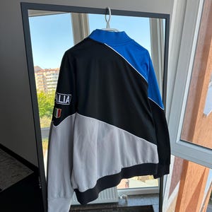 May include: A black, blue, and gray athletic jacket with the word "ITALIA" on the sleeve. The jacket has a white stripe design and a high collar. The Italian flag is on the sleeve. The jacket is hanging on a hanger.