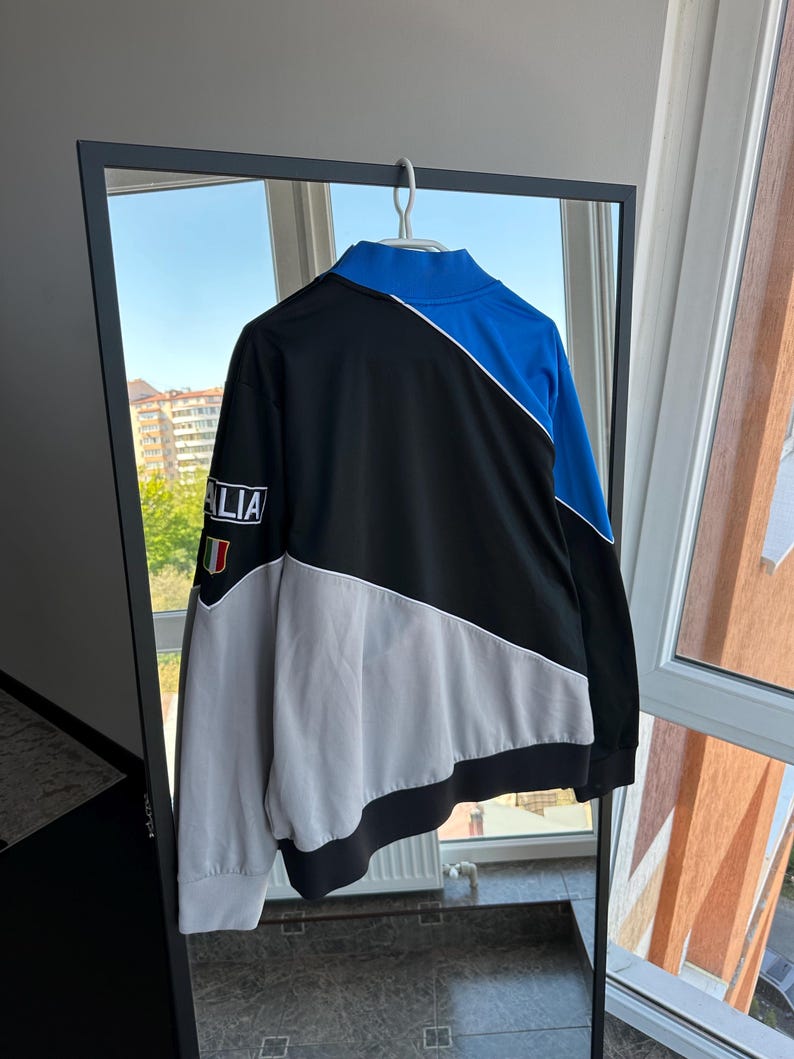 May include: A black, blue, and gray athletic jacket with the word "ITALIA" on the sleeve. The jacket has a white stripe design and a high collar. The Italian flag is on the sleeve. The jacket is hanging on a hanger.