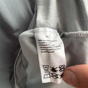 以下が含まれることがあります： 薄いグレーの衣服のクローズアップ。白いタグにはサイズ「XL」、生地の詳細、ケアマークが表示されています。生地は100％リヨセルです。