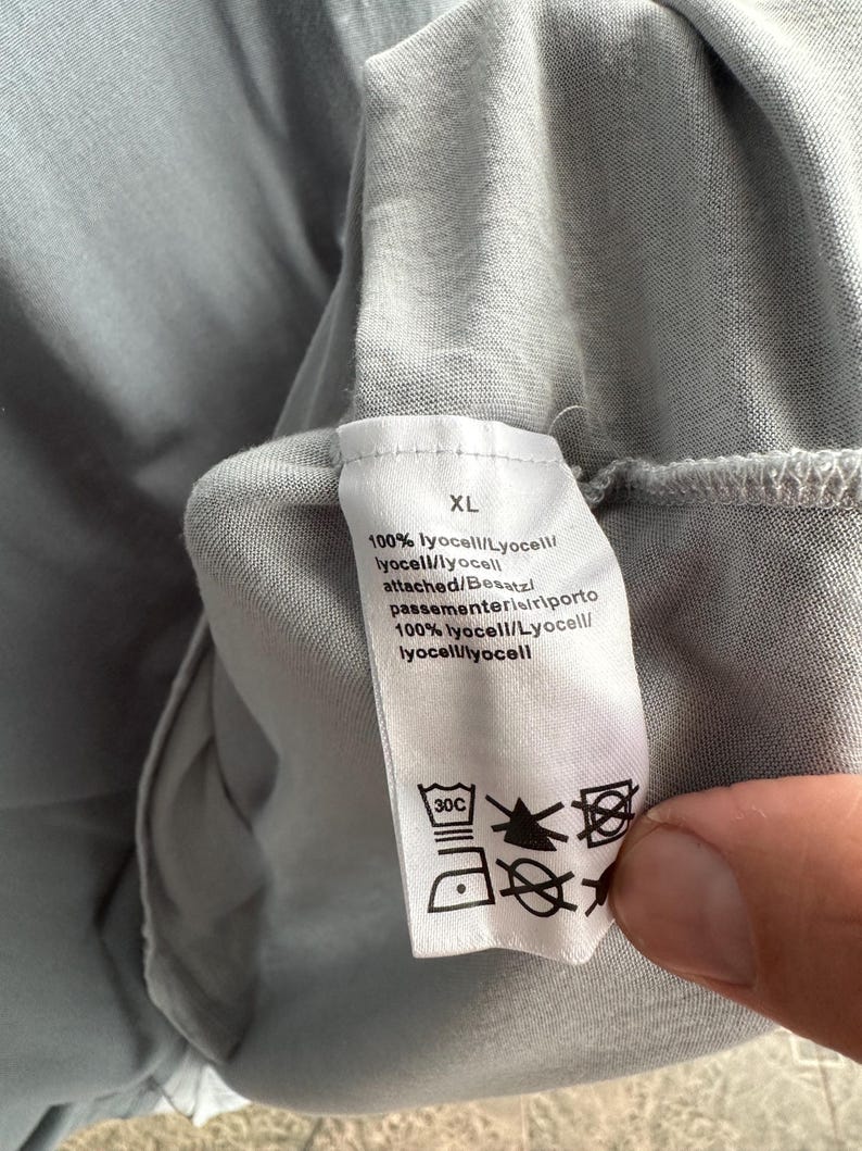 以下が含まれることがあります： 薄いグレーの衣服のクローズアップ。白いタグにはサイズ「XL」、生地の詳細、ケアマークが表示されています。生地は100％リヨセルです。