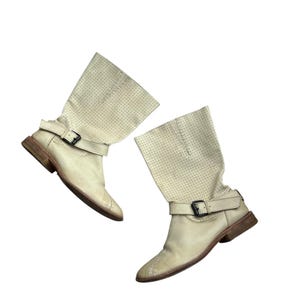 May include: A pair of cream-coloured leather boots with a perforated pattern on the upper shaft. Each boot has a buckle strap around the ankle and a brown sole. The boots have a small heel and a pointed toe.