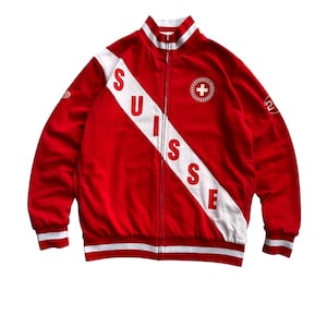 Puede incluir: Chaqueta deportiva roja con detalles blancos y cremallera completa. La chaqueta presenta una franja diagonal blanca con la palabra "SUISSE" en letras rojas. Un emblema de la cruz suiza está en el pecho.