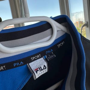 May include: A blue, black, and white Fila Sport polo shirt with a white label. The shirt is hanging on a white hanger. The Fila logo is red and white. The shirt has a blue collar and a black and white striped design.