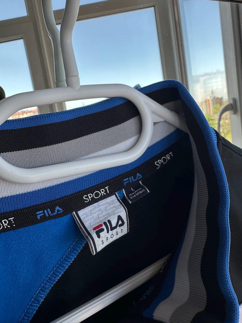 May include: A blue, black, and white Fila Sport polo shirt with a white label. The shirt is hanging on a white hanger. The Fila logo is red and white. The shirt has a blue collar and a black and white striped design.
