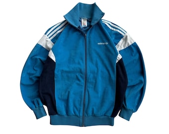 May include: A vintage blue Adidas track jacket with white stripes on the sleeves and shoulders. The jacket has a full zipper, a high collar, and the Adidas logo embroidered on the chest. The jacket has a blue, white, and black color block design.