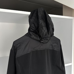 K&ouml;nnte beinhalten: Schwarze Kapuzenjacke mit Kordelzug. Die Jacke hat einen kontrastierenden Einsatz &uuml;ber den Schultern und einen durchgehenden Rei&szlig;verschluss. Die Kapuze ist gesteppt, und der K&ouml;rper der Jacke scheint aus glattem, mattem Stoff zu sein.