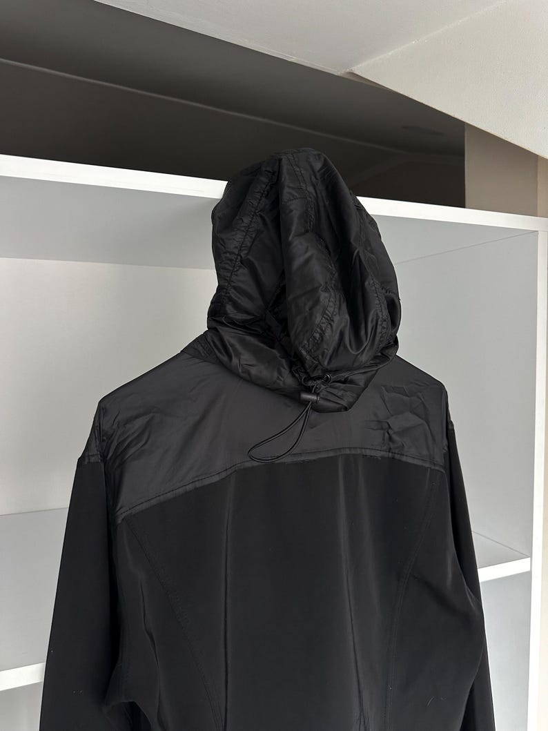 K&ouml;nnte beinhalten: Schwarze Kapuzenjacke mit Kordelzug. Die Jacke hat einen kontrastierenden Einsatz &uuml;ber den Schultern und einen durchgehenden Rei&szlig;verschluss. Die Kapuze ist gesteppt, und der K&ouml;rper der Jacke scheint aus glattem, mattem Stoff zu sein.