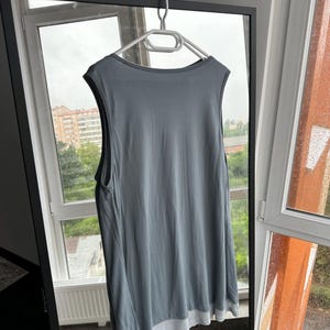以下が含まれることがあります： 白いハンガーにかけられた、白い裾が付いたスレートブルーのノースリーブトップ。 トップは柔らかく流れるような生地で作られています。 衣類は鏡の前に展示されており、窓と屋外の風景が映っています。