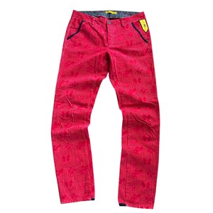 Op de afbeelding: Felrode broek met een geometrisch bloemmotief. De broek heeft een slanke pasvorm, met marineblauwe details op de zakken en tailleband. Een geel label is aan de zijkant bevestigd.