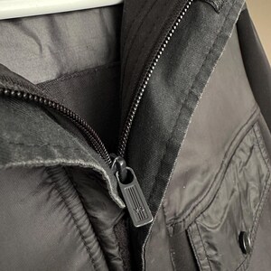 K&ouml;nnte beinhalten: Nahaufnahme einer schwarzen Jacke mit Rei&szlig;verschluss und zwei Taschen. Der Rei&szlig;verschluss hat ein rechteckiges Design. Die Jacke h&auml;ngt an einem wei&szlig;en Kleiderb&uuml;gel.