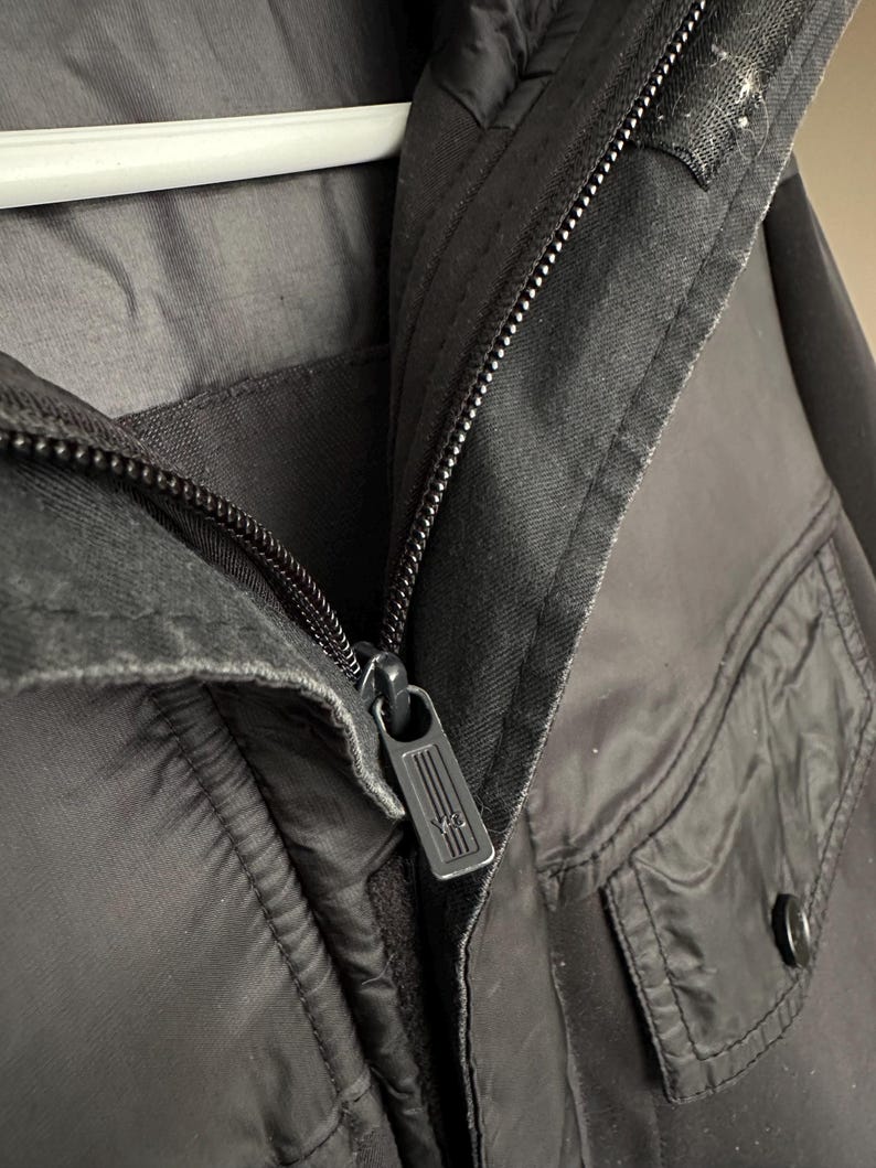 K&ouml;nnte beinhalten: Nahaufnahme einer schwarzen Jacke mit Rei&szlig;verschluss und zwei Taschen. Der Rei&szlig;verschluss hat ein rechteckiges Design. Die Jacke h&auml;ngt an einem wei&szlig;en Kleiderb&uuml;gel.