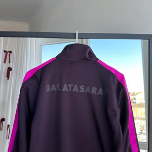May include: A dark purple Galatasaray track jacket with a high collar, hanging on a white hook. The jacket features bright pink accents on the shoulders and the text "GALATASARAY" across the back.
