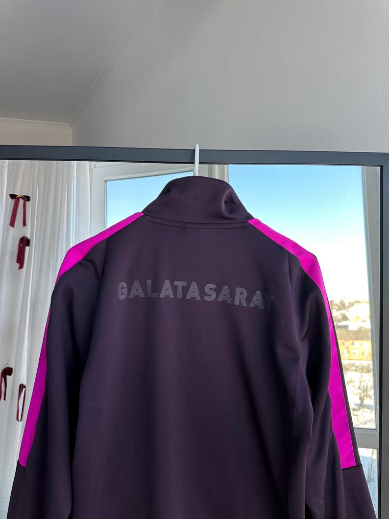 May include: A dark purple Galatasaray track jacket with a high collar, hanging on a white hook. The jacket features bright pink accents on the shoulders and the text "GALATASARAY" across the back.