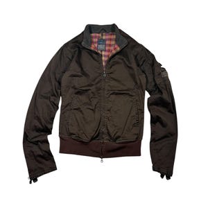 Puede incluir: Chaqueta marrón con cremallera y dobladillo y puños acanalados marrón oscuro. La chaqueta tiene un cuello gris y un forro a cuadros en tonos rosa, rojo y amarillo. Se ve un pequeño parche en la manga superior izquierda.