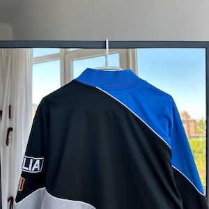 May include: A black, blue, and white track jacket hanging on a white hanger. The jacket has a high collar and a color-blocked design. The left sleeve features the word "LIA" and a small flag.