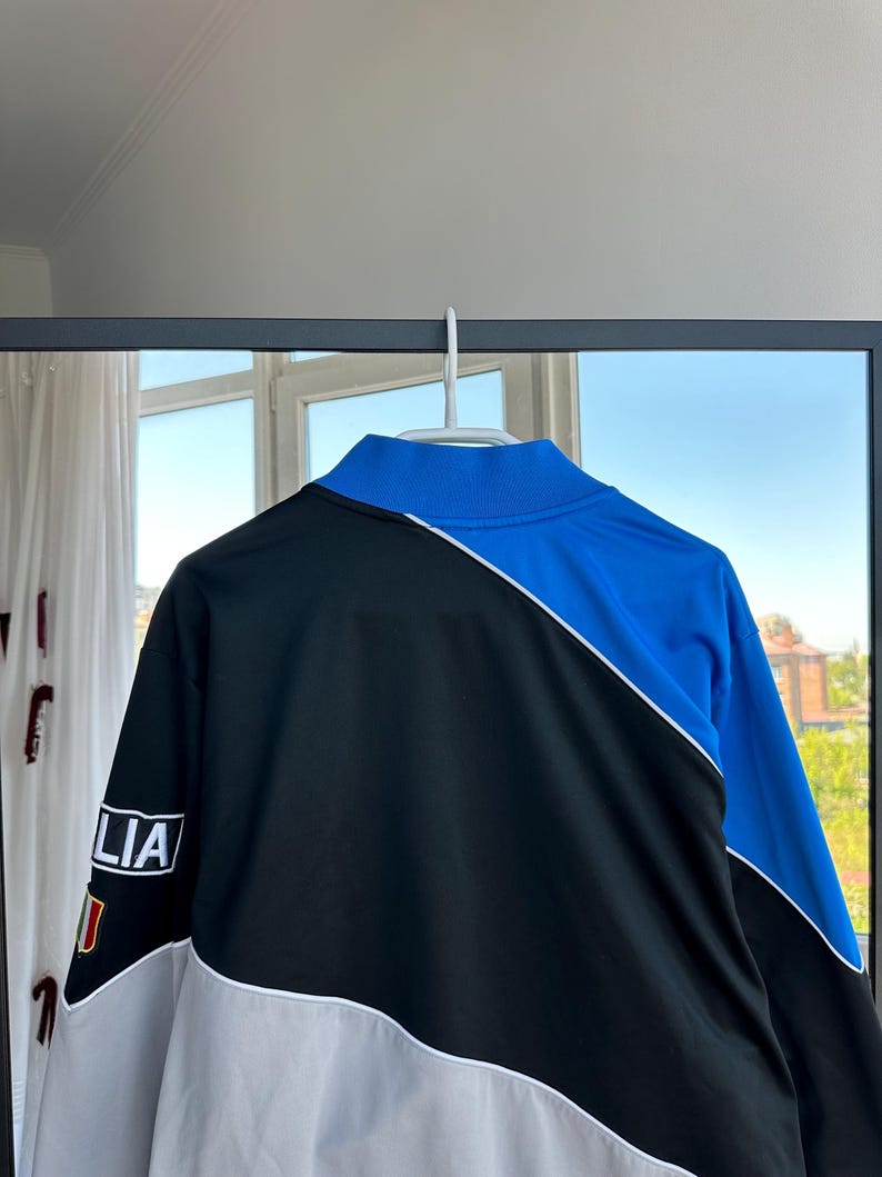 May include: A black, blue, and white track jacket hanging on a white hanger. The jacket has a high collar and a color-blocked design. The left sleeve features the word "LIA" and a small flag.